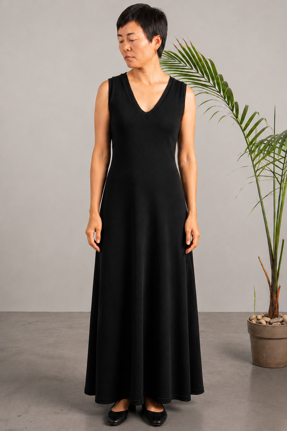 A woman modeling a sleeveless black maxi dress with a V-neckline, standing in a studio setting next to a potted palm tree.